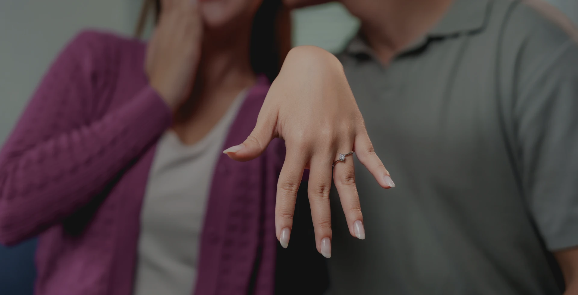 A woman's hand is held out to display an engagement ring. She is smiling, with a hand covering her mouth