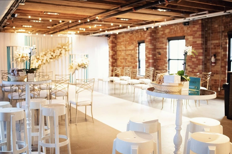 A brick and wooden loft decorated with white chairs and tables and soft yellow lighting.