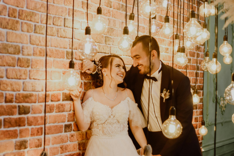 Couple in wedding attire smiling at each other next to an exposed brick wall with light bulbs around them 